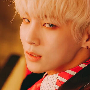 a screencap of key from shinee in his bad love music video. he has blond hair and red eye makeup; he is smoldering at the camera in a dark suit with a white/red shirt, because he's a menace.